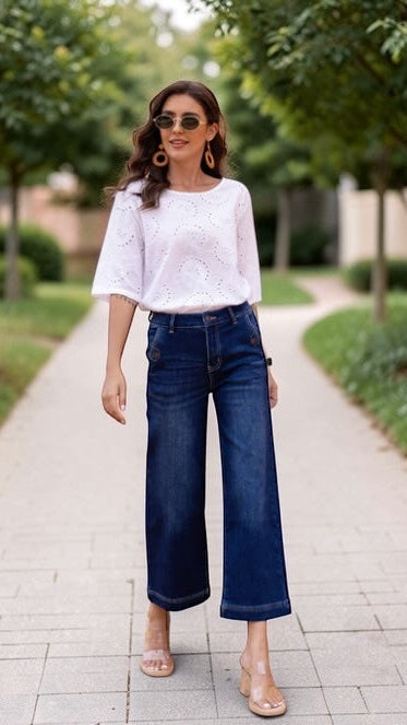 Woman wearing a white blouse and blue jeans standing on a paved path with greenery in the background