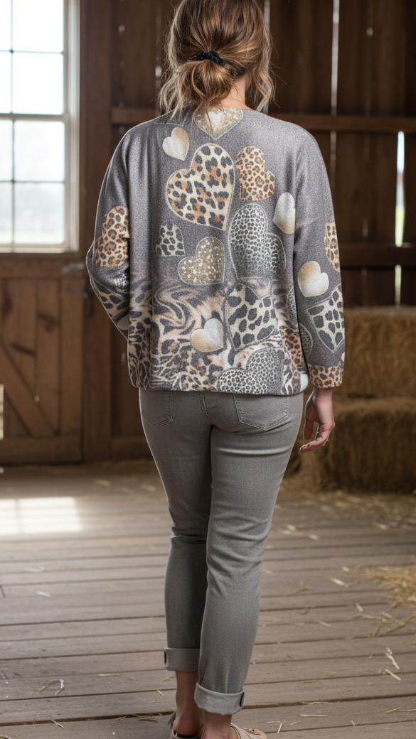 A woman viewed from behind wearing a gray long-sleeve cardigan with a mixed abstract and animal-print pattern, paired with gray pants and tan slide sandals, standing on a wooden barn floor with hay bales in the background.